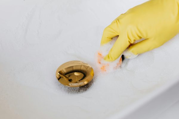 From above of crop anonymous person in yellow rubber protective glove using sponge and detergent for washing white sink in bathroom Vue de dessus d'une personne anonyme en gant de protection en caoutchouc jaune utilisant une éponge et un détergent pour laver un lavabo blanc dans la salle de bain.