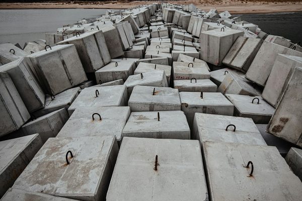 Stacked concrete blocks line a beach under an overcast sky, suggesting a moody atmosphere. Des blocs de béton empilés bordent une plage sous un ciel couvert, suggérant une atmosphère morose.