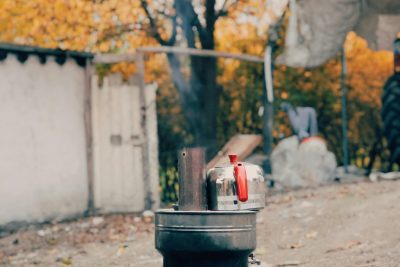 A rustic outdoor stove with a kettle boiling, surrounded by autumn foliage. Un poêle extérieur rustique avec une bouilloire qui bout, entouré de feuillage automnal.