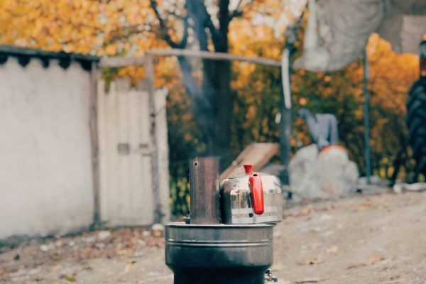 A rustic outdoor stove with a kettle boiling, surrounded by autumn foliage. Un poêle extérieur rustique avec une bouilloire qui bout, entouré de feuillage automnal.