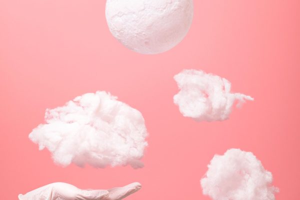 An artistic composition of floating clouds and hand with medical glove against a pink backdrop. Une composition artistique de nuages flottants et d'une main avec un gant médical sur un fond rose.