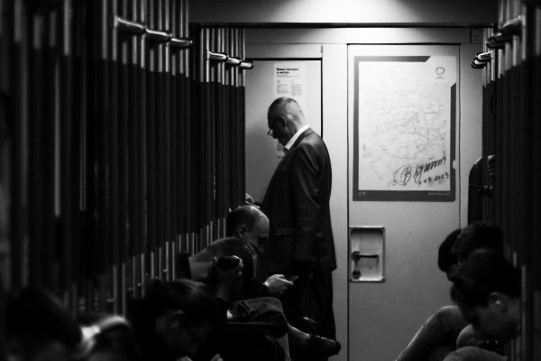 Black and white image of people sitting and standing in a train corridor, using smartphones. Image en noir et blanc de personnes assises et debout dans un couloir de train, utilisant des smartphones.