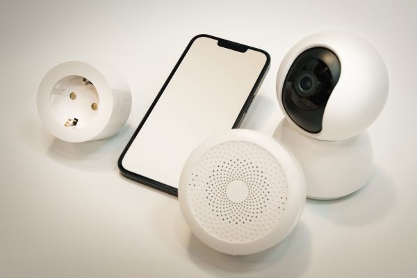 Smart home gadgets including a security camera, speaker, and smartphone displayed on a white surface. Gadgets de maison intelligente incluant une caméra de sécurité, un haut-parleur et un smartphone affichés sur une surface blanche.